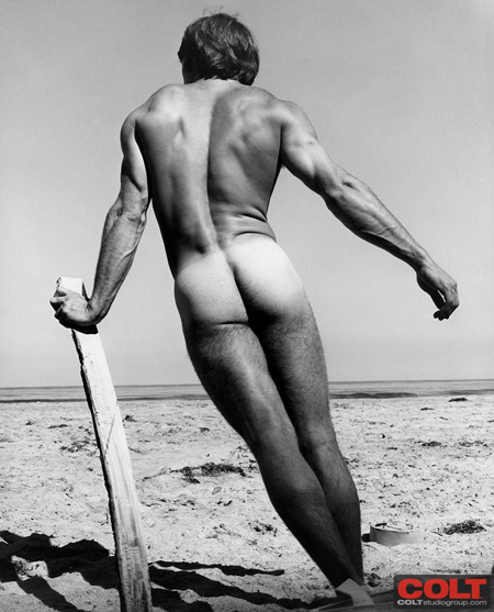 Classic black and white erotic photo of a muscular young man nude at the ocean with his tan lines exposed.